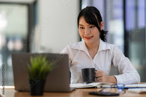 Attractive businesswoman holding cup of hot drink and looking away with smile while sitting in office.