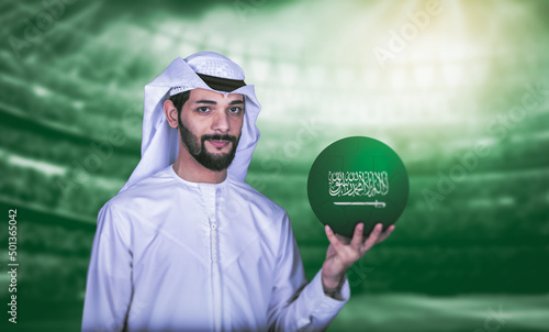 Arab man holding Saudi soccer ball standing front green stadium background.