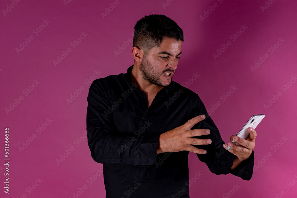 A man with a black shirt, posing on a pink background looking at the cell phone with a gesture of anger and fury. Emotions, joy and fun of men. A man with a black shirt, posing on a pink background looking at the cell phone with a gesture of anger and fury. Emotions, joy and fun of men.