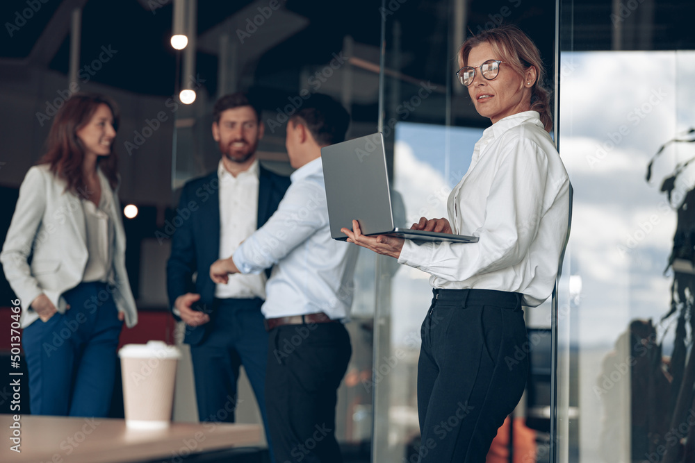 Mature confident businesswoman at office with group of colleagues on background, working on laptop. Teamwork and business concept Mature confident businesswoman at office with group of colleagues on background, working on laptop. Teamwork and business concept