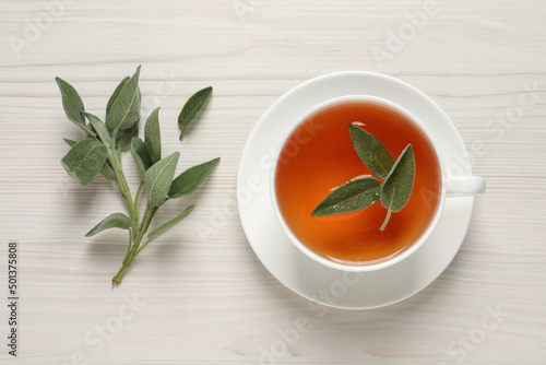 Cup of aromatic sage tea and fresh leaves on white wooden table, flat lay