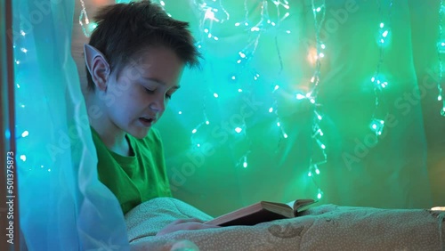 a teenager with long ears reads a book sitting on a bed decorated with garlands. wrapped in a blanket. elf costume