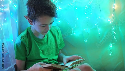 a teenager in an elf costume reads a book sitting on a bed in the light of garlands. fabulous light from the lights. a boy in a fairy tale