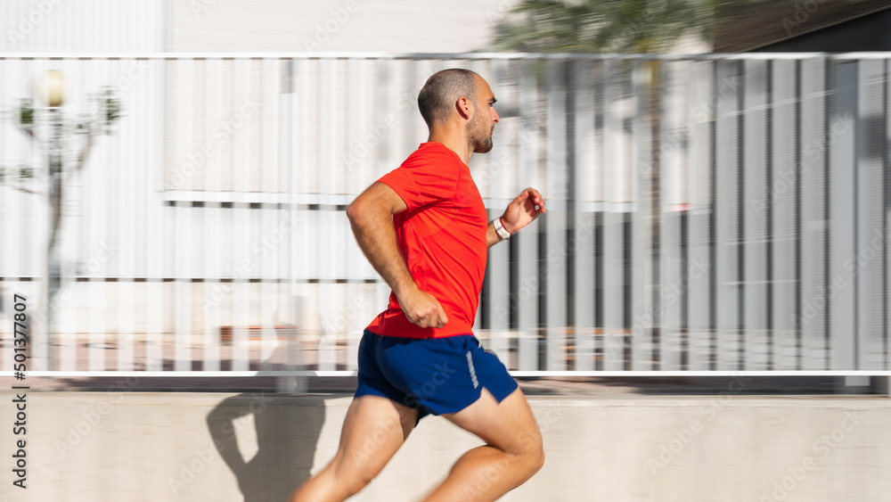 Caucasian man running on a sunny day. He is wearing a red T-shirt and blue shorts. Background with motion blur. Spain, Alicante Caucasian man running on a sunny day. He is wearing a red T-shirt and blue shorts. Background with motion blur. Spain, Alicante