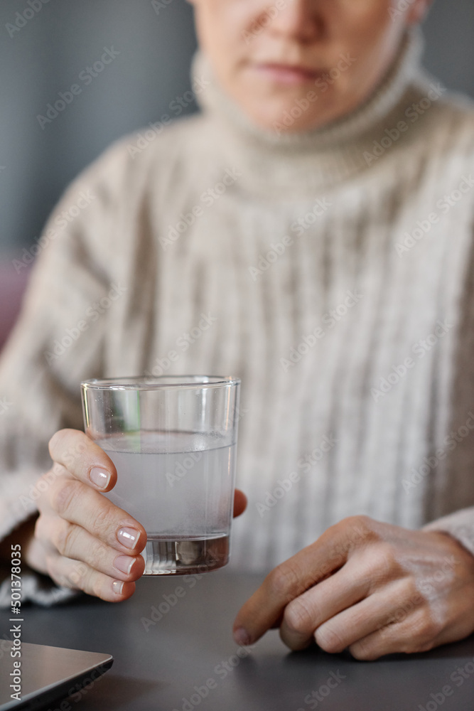 Vertical shot of mature woman having cold wearing sweater sitting at desk at home holding glass with mixture of water and effervescent tablet Vertical shot of mature woman having cold wearing sweater sitting at desk at home holding glass with mixture of water and effervescent tablet
