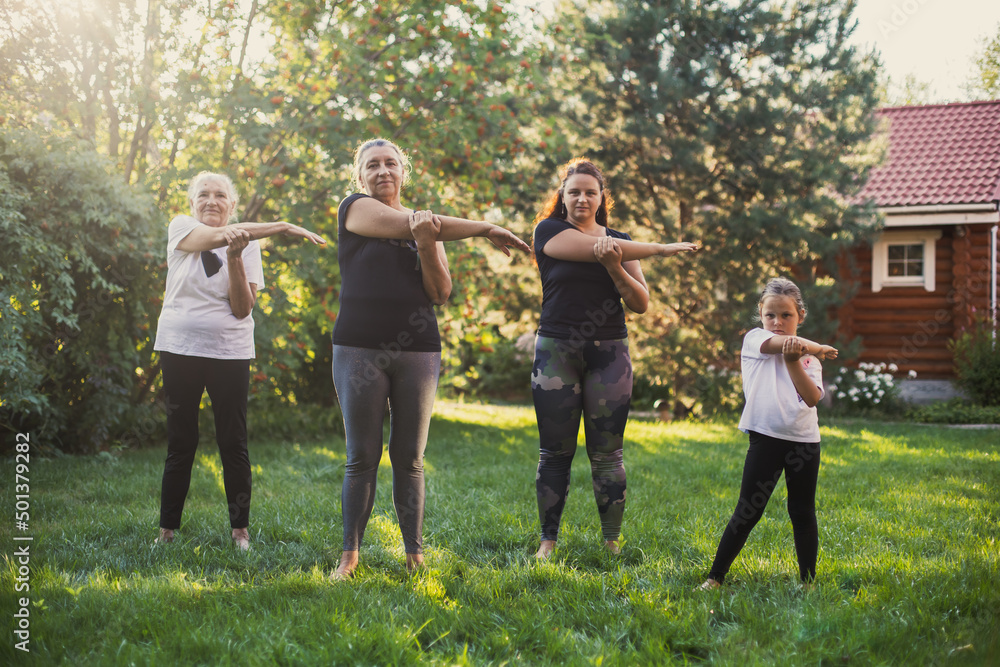 Females of few generations exercising outside stretching arms together in same way standing on meadow full of green grass and trees. Spending time together. Females of few generations exercising outside stretching arms together in same way standing on meadow full of green grass and trees. Spending time together.