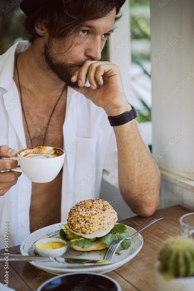 Young handsome man in a white shirt open, having breakfast in a cafe with a vegetarian burger, drinking coffee, lifestyle in a tropical island, life in Bali.