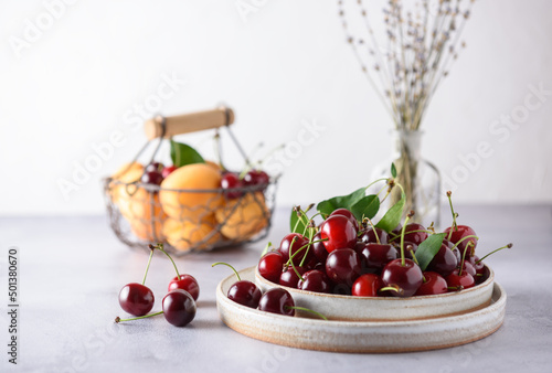 Fresh organic cherries in a grey ceramic plate on a light grey background. In the background there is a basket with apricots and cherries and a saucer with cherries. Upper angle.