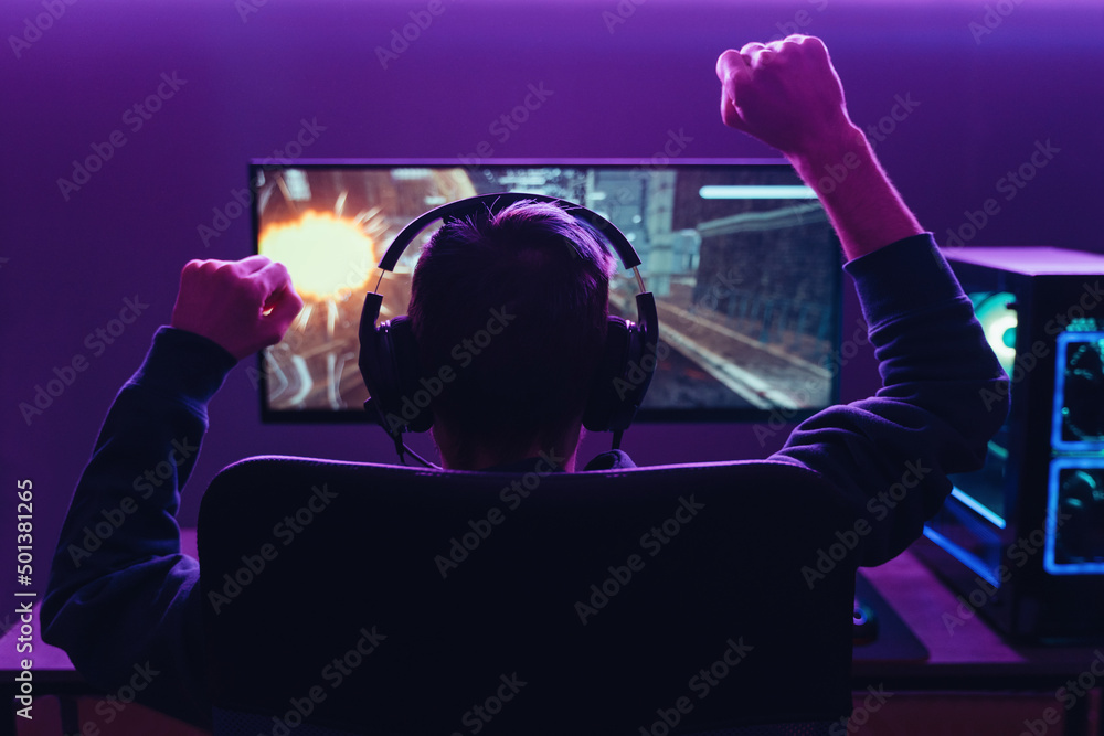 Back view of pro gamer wearing headset celebrating victory in online internet esport tournament, makes yes hands gesture, looking at monitor. Neon coloured dark room. Cyber sport and e-sport concept - obrazy, fototapety, plakaty 