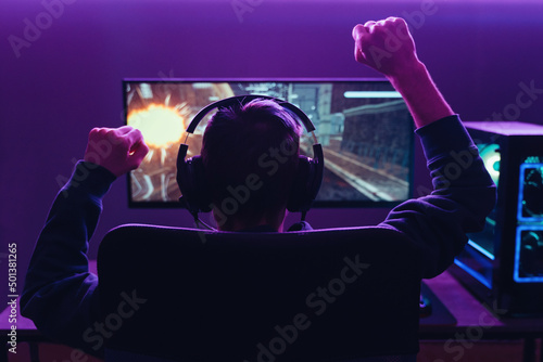Back view of pro gamer wearing headset celebrating victory in online internet esport tournament, makes yes hands gesture, looking at monitor. Neon coloured dark room. Cyber sport and e-sport concept photo