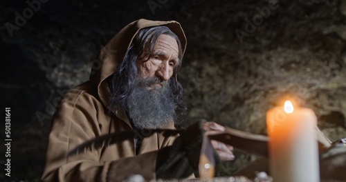 Elderly monk taking notes with quill in cave