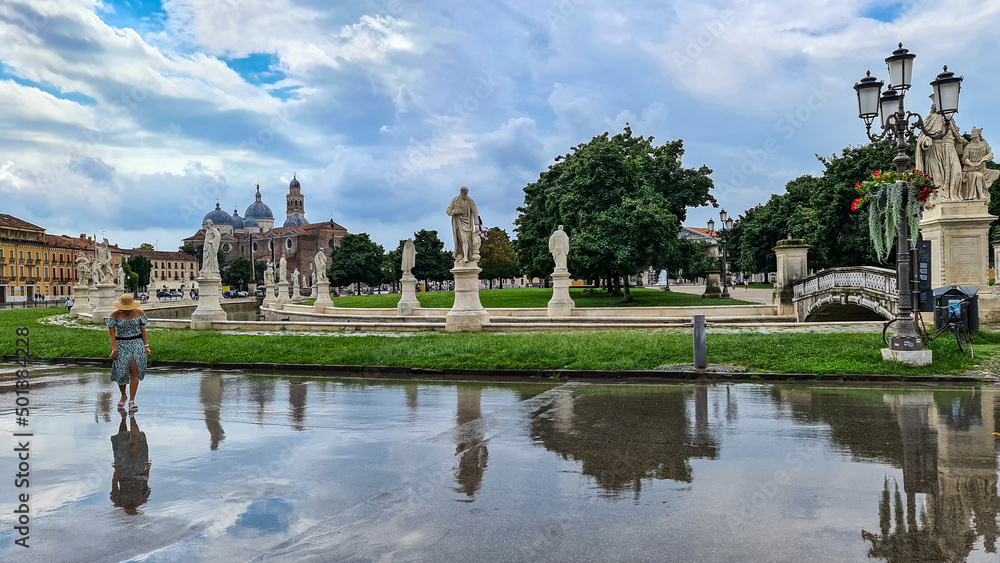 Woman walking on the center square after rain. Scenic view after strong rain on Prato della Valle, Abbey of Santa Giustina, city of Padua, Veneto, Italy, Europe. Water on street creates reflection
