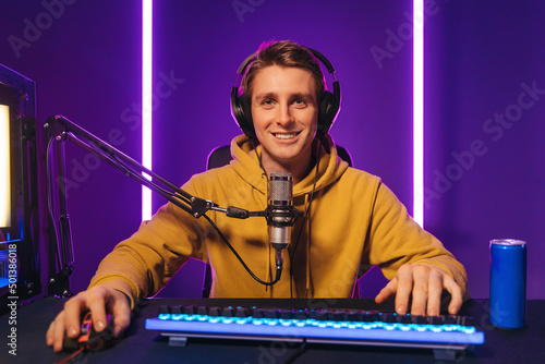 Smiling professional gamer streaming esport tournament, playing online video game at home, using microphone and headphones, drinking energy caffeine drink, enjoying the game, looking at camera 