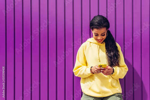 Young woman of african-american ethnicity using her smatphone on purple background. Influencer girl with yellow clothes using her mobile phone and her social networks. photo