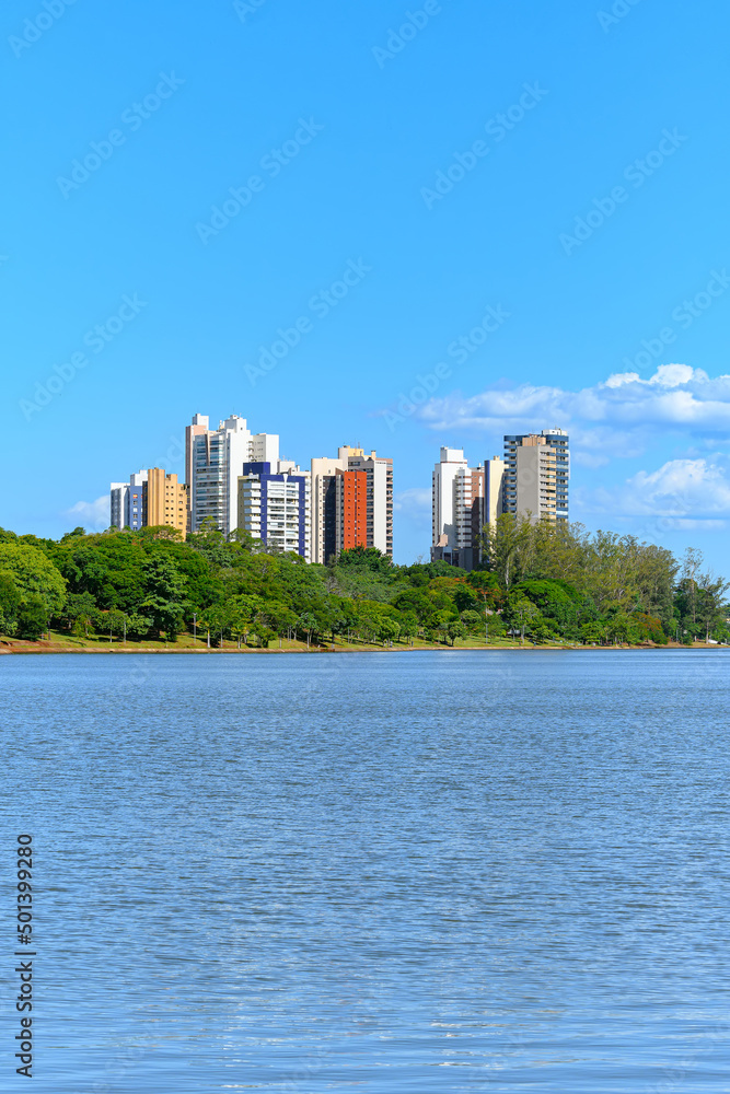 Landscape of Igapo lake, Londrina - PR, Brazil. Beautiful city lake, with trees around the shore and the city buildings on background, on a blue sky day.