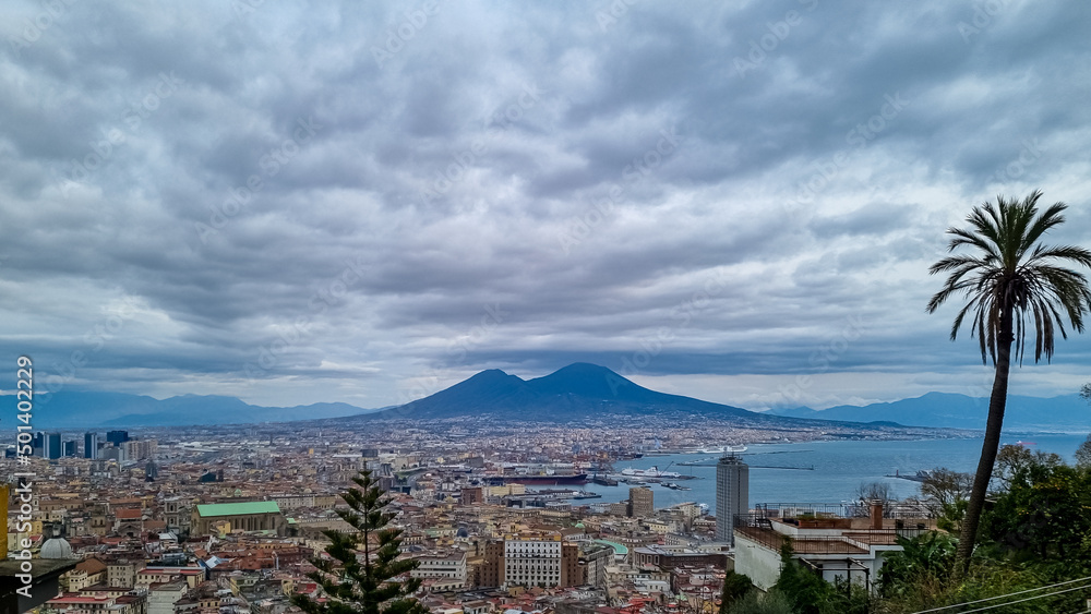 Fototapeta premium Panoramic view from Vomero on volcano Mount Vesuvius in Naples, Campania, Italy, Europe. Ferries in the port of Naples. Clouds are accumulating around the mountains. Palm tree with sea view
