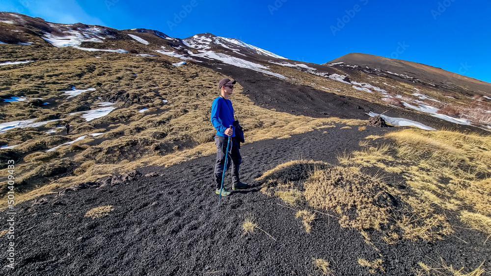Man surrounded by golden grass hiking on volcanic landscape of volcano mount Etna, in Sicily, Italy, Europe. Barren terrain of solidified lava, ash and pumice. Slopes of crater covered with snow