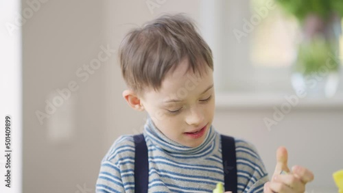 Joyful smiling autistic boy painting with colors sitting at table indoors. Front view portrait of Caucasian kid with mental disorder studying at special school. Education and individuality concept photo