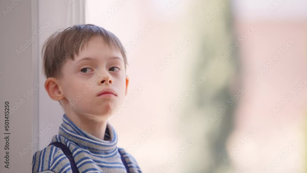 Caucasian autistic boy standing at window indoors looking away. Portrait of child with mental disorder at home on sunny day. Lifestyle and autism concept Caucasian autistic boy standing at window indoors looking away. Portrait of child with mental disorder at home on sunny day. Lifestyle and autism concept