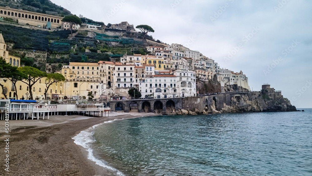 Scenic view from the beach near the harbor at the coastal village of Amalfi at Mediterranean Sea in Campania, Italy, Europe. Warm sunlight is touching the waterfront houses. Golden hour, Amalfi Coast