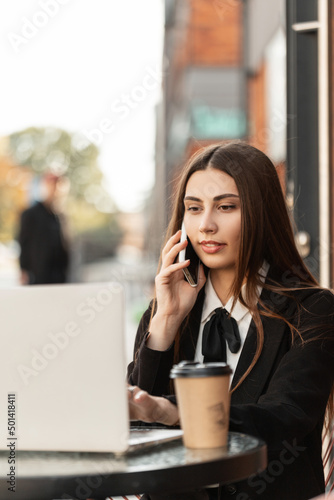 Happy beautiful hr manager woman in a fancy black suit with a tie and shirt works on a laptop and talks on the phone, sitting and drinking coffee in a cafe in town