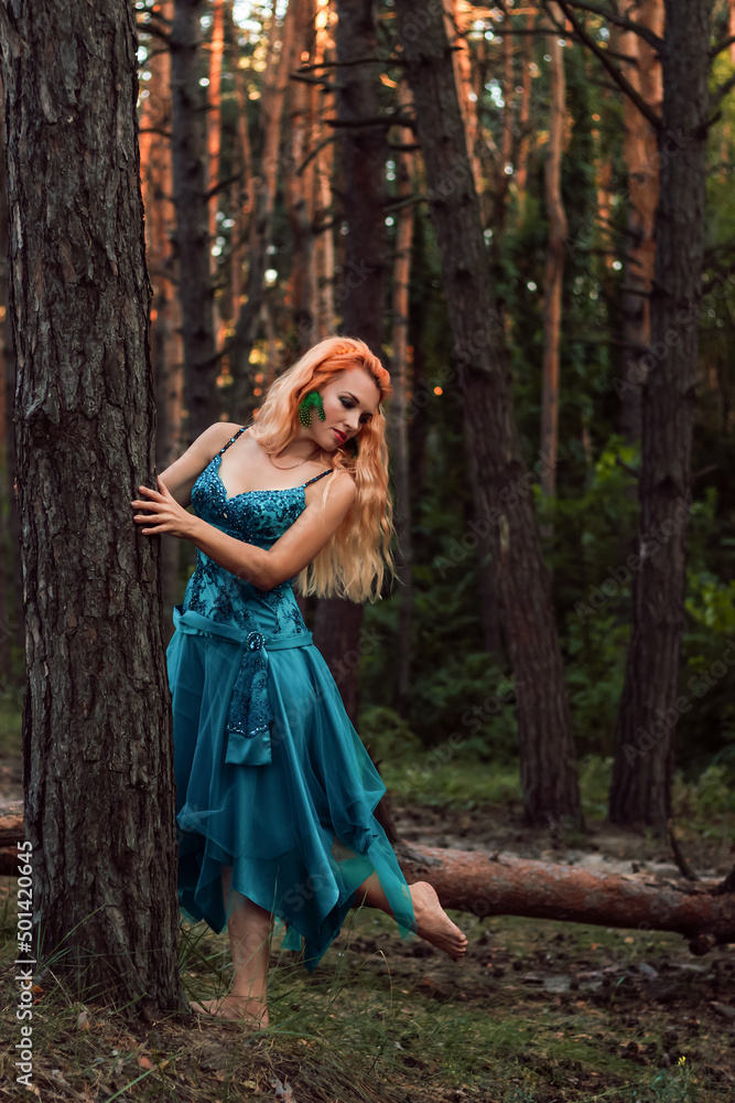 A beautiful girl with red hair stands near a tree in the forest and looks away A beautiful girl with red hair stands near a tree in the forest and looks away