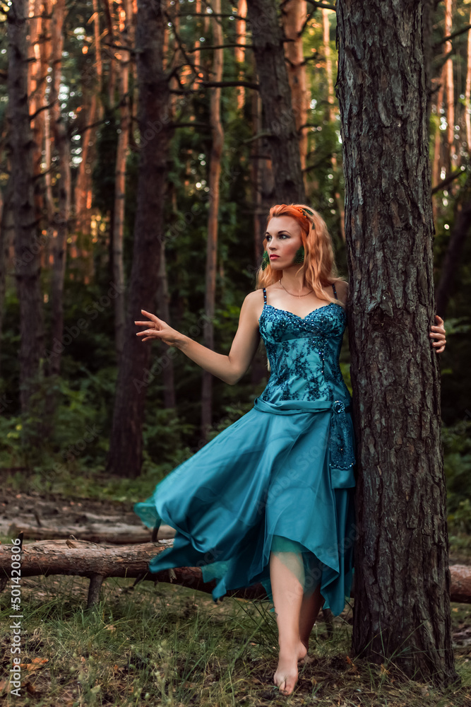 A beautiful girl with red hair stands near a tree in the forest and looks away