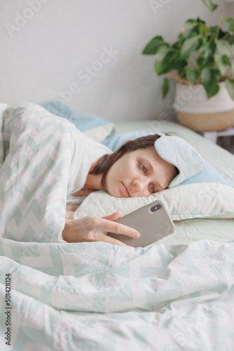 woman in a sleep mask is lying in bed and chatting on a smartphone. a European woman with sleep problems and insomnia, waking up and falling asleep in a cozy house. sleep disorders or depression