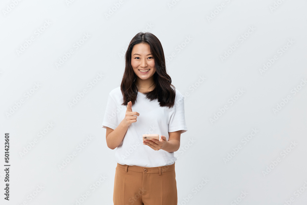 Happy smiling cute Asian young female in white basic t-shirt typing message chatting with boyfriend using phone posing isolated on white studio background. The best offer for ad. Social media concept Happy smiling cute Asian young female in white basic t-shirt typing message chatting with boyfriend using phone posing isolated on white studio background. The best offer for ad. Social media concept