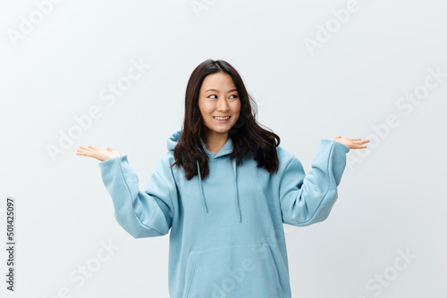 Cheerful happy cute Asian young female in blue hoodie sweatshirt holds invisible objects on palms posing isolated on white studio background. The best offer for ad. Fashion New Collection Ad concept