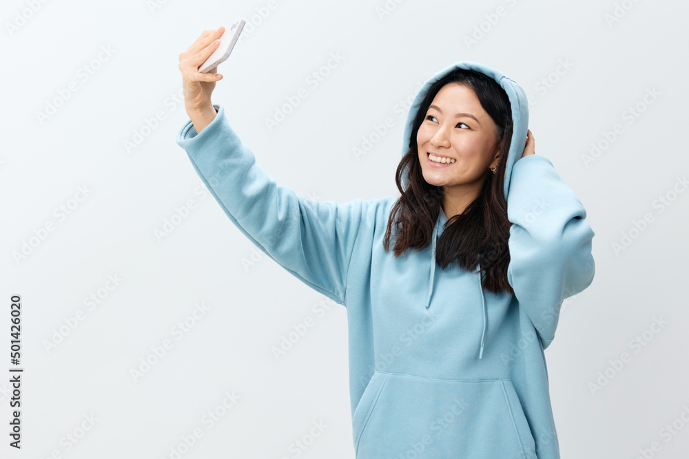 Smiling pretty Korean young female in blue hoodie hold phone doing selfie video call posing isolated on over white studio background. The best offer for ad. Distance communication Gadget Ad concept Smiling pretty Korean young female in blue hoodie hold phone doing selfie video call posing isolated on over white studio background. The best offer for ad. Distance communication Gadget Ad concept