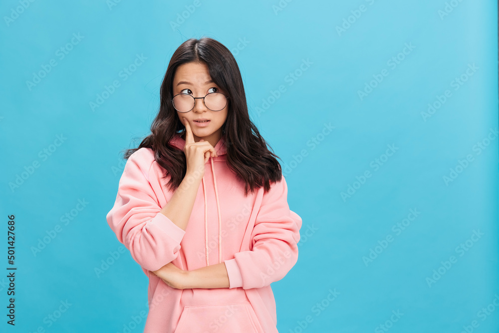 Pensive Asian young lady in pink hoodie sweatshirt wear round glasses can not make choose posing isolated on over blue studio background. The best offer for ad. Eyewear for vision correction concept Pensive Asian young lady in pink hoodie sweatshirt wear round glasses can not make choose posing isolated on over blue studio background. The best offer for ad. Eyewear for vision correction concept