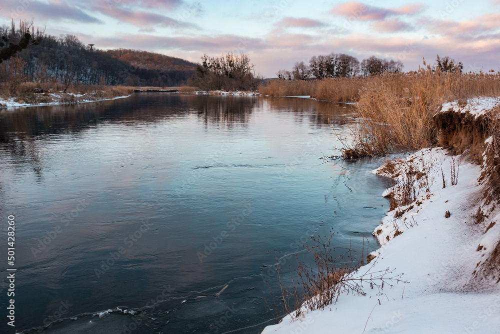 Fototapeta premium Winter view on cold river with scenic colorful clouds with reflection in mirror water and dry reed grass on shore. Zmiyevsky region on Siverskyi Donets River covered in snow in Ukraine