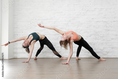 Two young women practicing stretching yoga positions in yoga studio. Wellbeing and self care concept. 