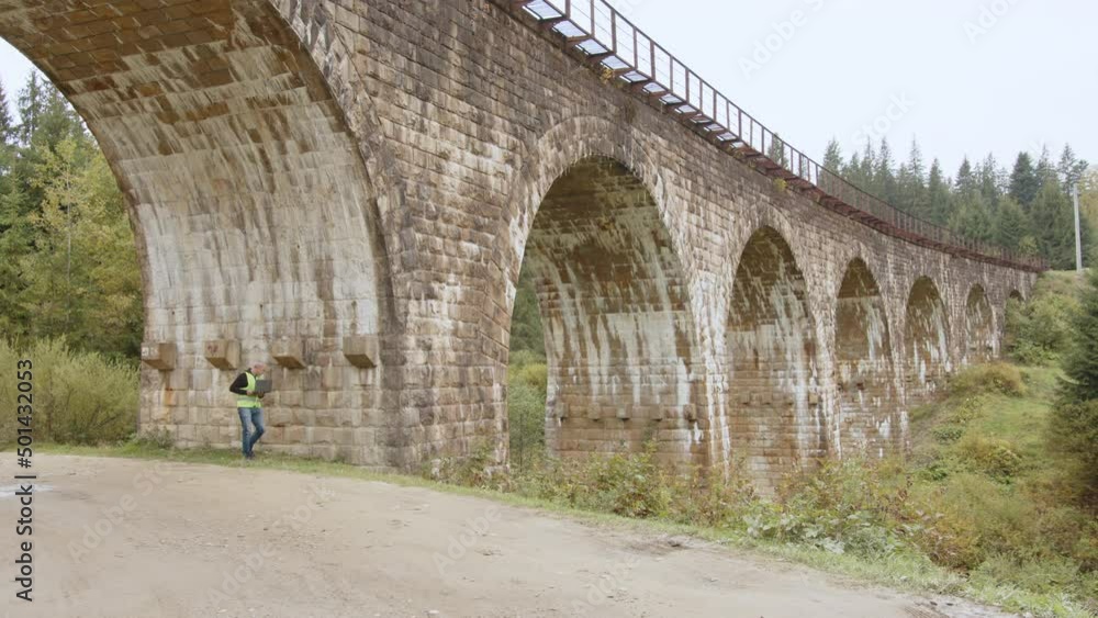 Senior male engineer to restore ancient buildings or bridges. Engineer writes data to laptop. Reconstruction of bridges, preservation of ancient railway bridges. Engineer-inspector at work Senior male engineer to restore ancient buildings or bridges. Engineer writes data to laptop. Reconstruction of bridges, preservation of ancient railway bridges. Engineer-inspector at work