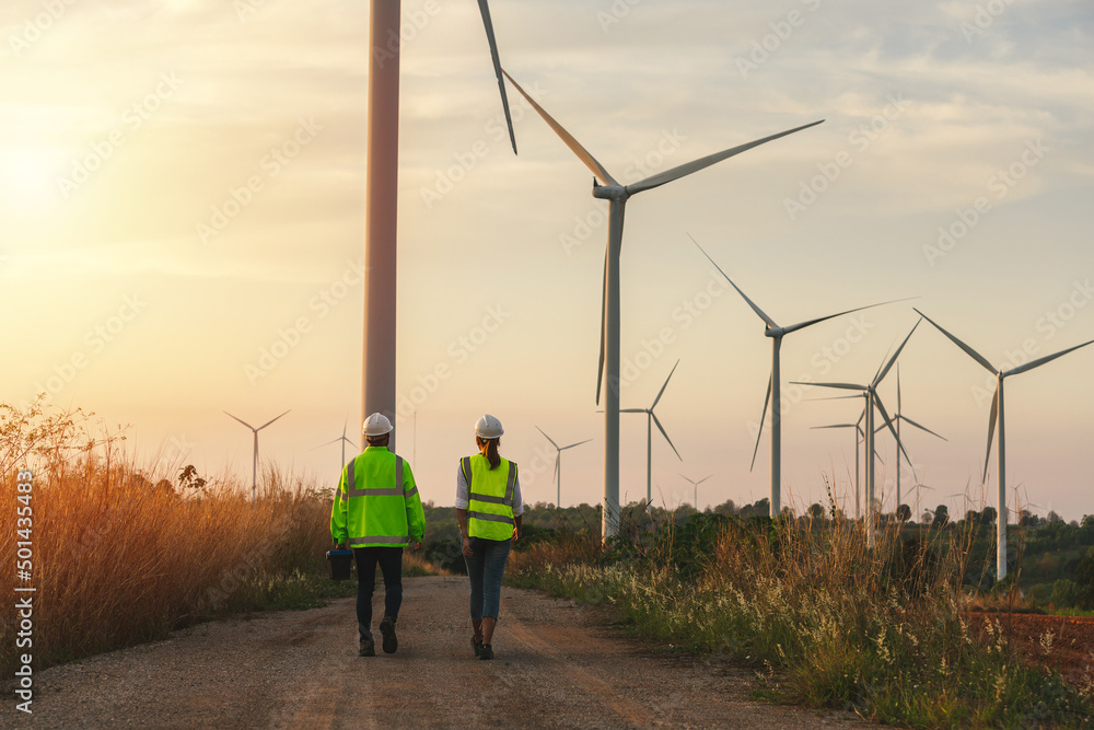 Obraz premium Back view of young maintenance engineers team working in wind turbine farm at sunset.
