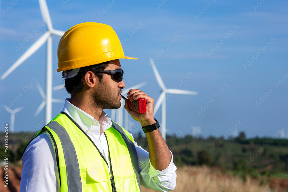 Young engineer wear protective helmet using radio communication in wind turbine farm on background. Young engineer wear protective helmet using radio communication in wind turbine farm on background.
