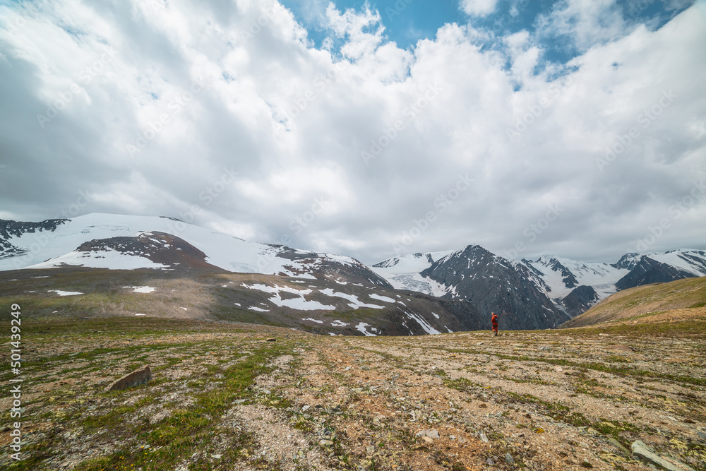 One man walks with view to high snowy mountain range with sharp tops and glaciers under gray cloudy sky. Colorful landscape with tourist on background of large snow mountains at changeable weather. One man walks with view to high snowy mountain range with sharp tops and glaciers under gray cloudy sky. Colorful landscape with tourist on background of large snow mountains at changeable weather.