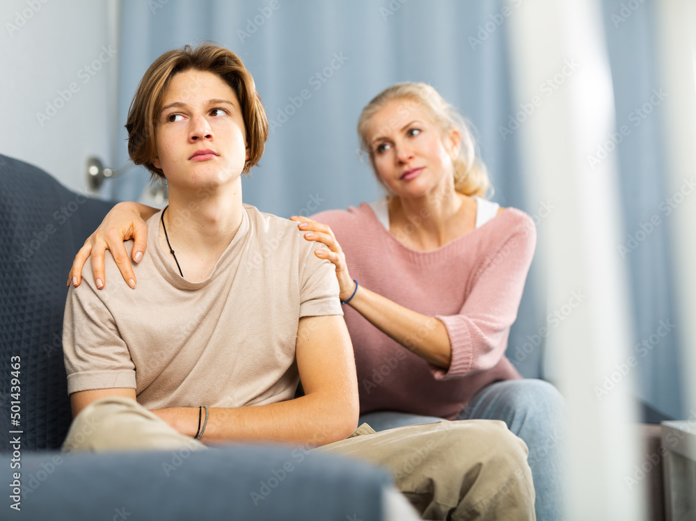 Obraz premium Friendly caring woman calming sad teenage son after disagreements, while sitting together in cozy living room..