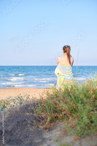 Summertime scenes. Unrecognizable young adult woman wrapped in a towel stands next to the beach shore looking into the ocean on a summer sunny day.