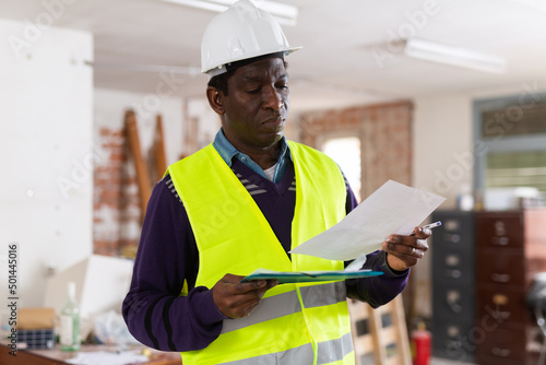 Strict focused african american building inspector wearing yellow safety vest and hard hat standing with papers at construction site indoors, checking safety and compliance with regulations..