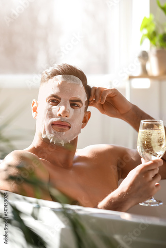 Foto Handsome man with facial mask and glass of wine taking bath at home