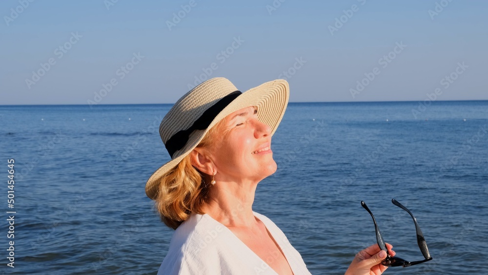 happy 50 year old woman in straw hat and sunglasses enjoying the sun against the blue sea background. Summer, vacation, vacation, active retirees