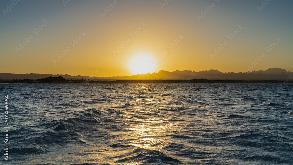 Sunset in Egypt. The sun is low over the mountain range. The sky is colored orange. Small waves on the Red Sea. A sunny path on the water. Safaga