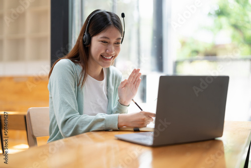 Smiling girl student wear wireless headphone study online with teacher, happy young asian woman learn language listen lecture watch webinar write notes look at laptop sit in cafe, distant education