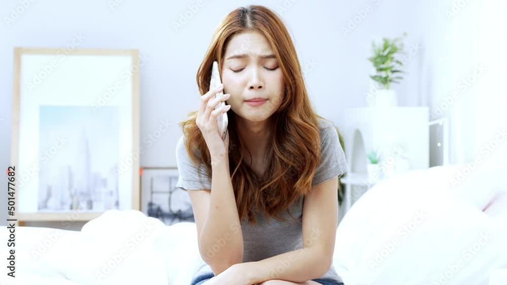 Young Asian woman sitting on bed talking seriously on smartphone in her bedroom, negative facial expression. Young Asian woman sitting on bed talking seriously on smartphone in her bedroom, negative facial expression.