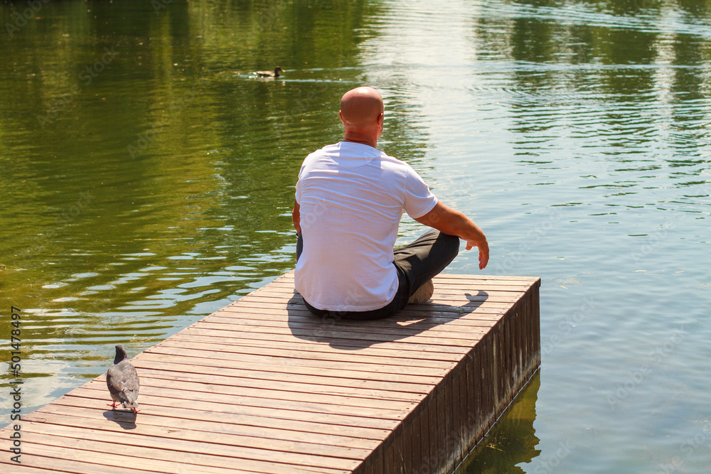 Lonely man sitting on edge wooden raft on pier looks out over lake on sunny day in the park. Solo travel through nature, wanderlust and inspirational concept Lonely man sitting on edge wooden raft on pier looks out over lake on sunny day in the park. Solo travel through nature, wanderlust and inspirational concept