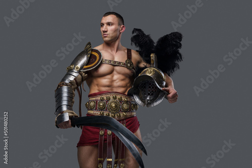 Studio portrait of gladiator athlete with naked torso holding sword and helmet against grey background.