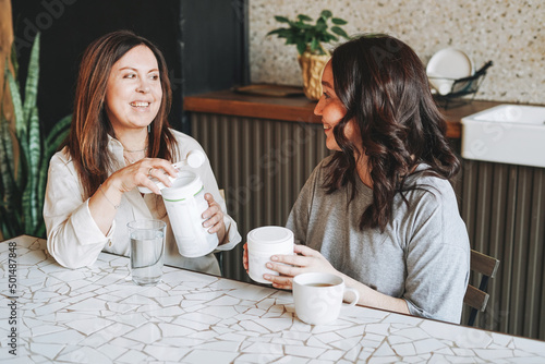 Adult smiling brunette middle aged womean having breakfast with dry instant protein vitamin supplement shake in kitchen at home photo