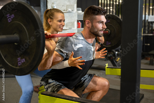 Muscular young sporty man lifting heavy weights with assistance of attractive blonde fit woman at gym